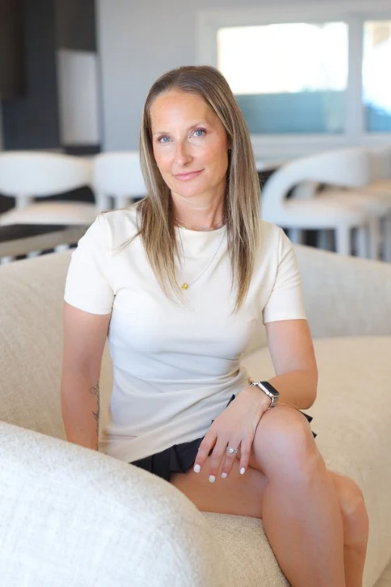 Sara Chiarilli, founder of Artful Conceptions, seated gracefully on a linen couch in a beautifully designed interior. She is dressed stylishly and smiling warmly, with a backdrop featuring soft, neutral tones, elegant throw pillows, and a curated side table, showcasing her expertise in creating cozy and sophisticated living spaces.