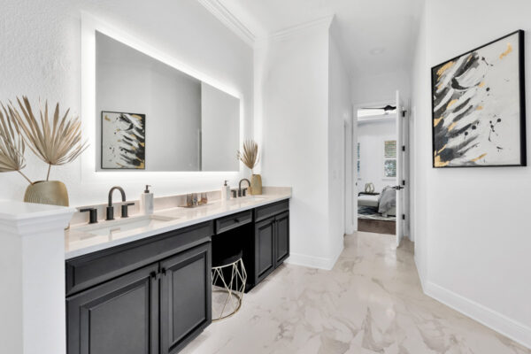 View from a bathroom leading into a spacious bedroom, with a modern design featuring light-colored walls and a minimalist aesthetic. The bathroom includes a sleek vanity and mirrors, while the bedroom beyond features soft, neutral tones and comfortable furnishings.