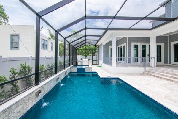 Beautiful new construction patio featuring a pool with decorative fountains. The pool is surrounded by a spacious stone patio, with lounge chairs placed around it for relaxation. Lush greenery and a clear blue sky enhance the tranquil outdoor setting.