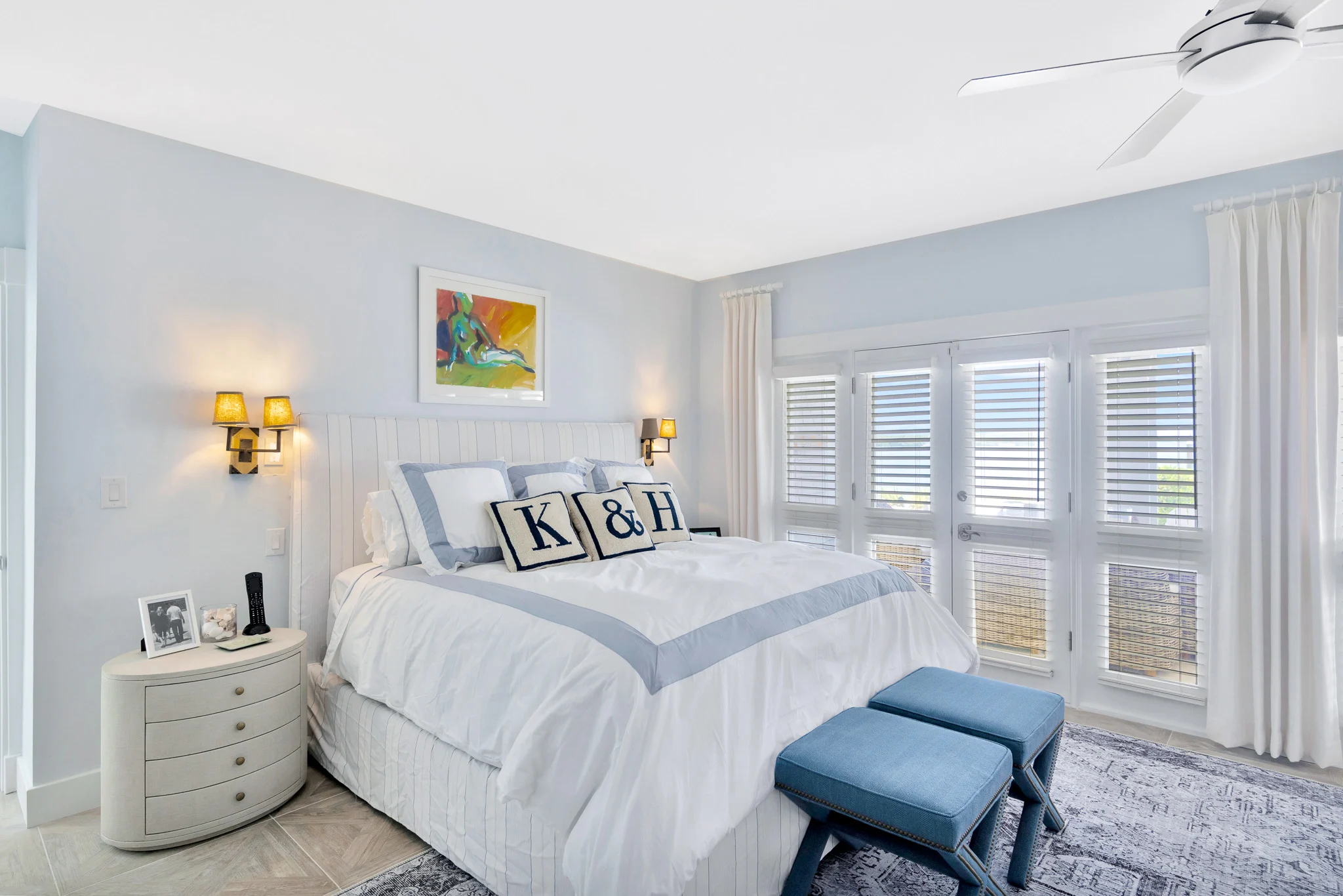 Coastal primary bedroom featuring custom upholstered bed, plantation shutters, soft blue accents, and waterfront views.